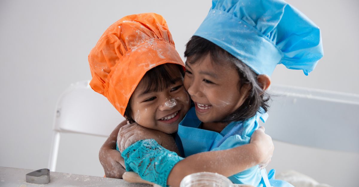 Two preschoolers wearing aprons and baking hats with flour on their faces as they smile and hug. 