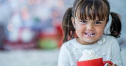 A preschool aged child smiling and drinking hot chocolate with whip cream.