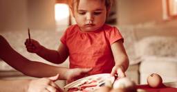 An infant sitting with their teacher looking at a tray of different paint colors