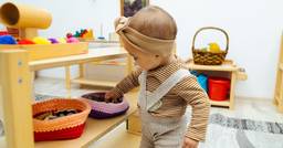 A young toddler exploring her classroom independently and feeling items on the shelves.