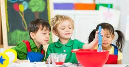 Three preschool children engaging in a science experiment as they use a whisk to mix items in a red bowl.