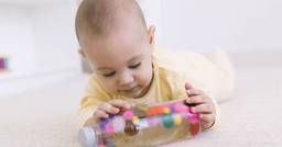 A young infant lying on their stomach as they explore a sensory bottle with colourful pom-poms inside of it