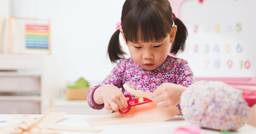 Preschooler practicing their fine motor skills as they cut paper with scissors at a table in the classroom.