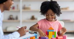 A kindergarten aged child smiling and sitting at a table building with colorful wooden blocks, with their teacher in the foreground taking notes.
