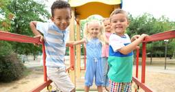 4 preschoolers smiling at the camera as they play on the playground outside